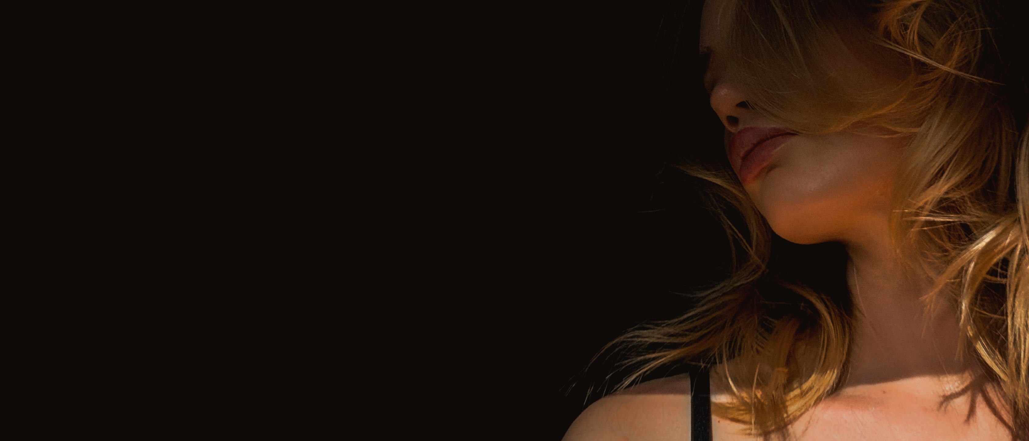 Close-up of lips, jaw and a bare shoulder, the face half hidden by windswept blonde hair in warm low light